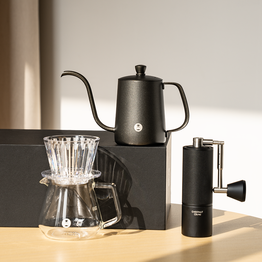 TIMEMORE C3S Pro black grinder alongside B75 transparent coffee dripper, glass server, and black pour-over kettle on wooden surface.